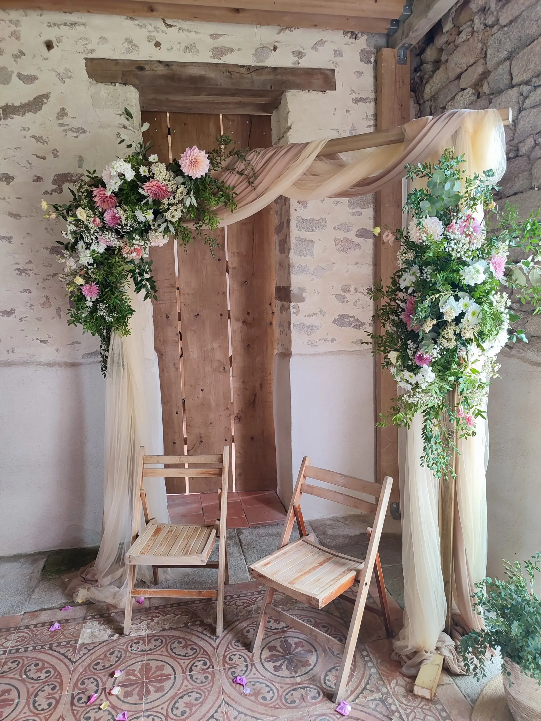 Arche de cérémonie laïque : fleurs roses et blanches, voilages et chaises en bois.