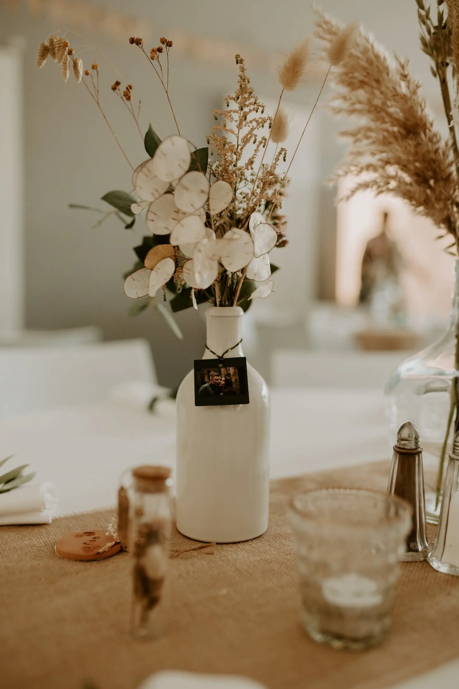 Bouquet de fleurs séchées et de monnaie-du-pape dans un vase blanc, sur une table de fête.