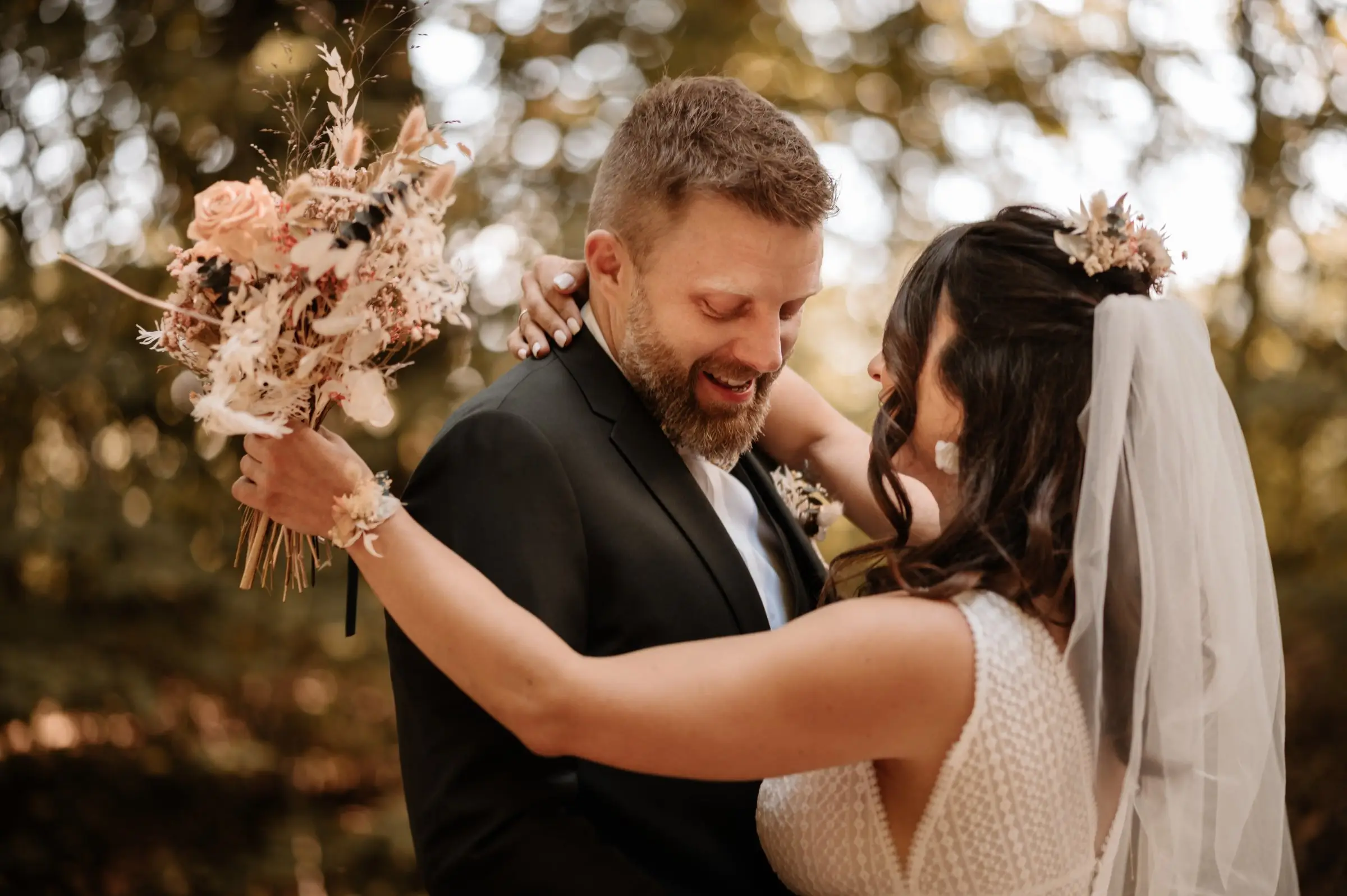 Une mariée enlace son mari en levant un bouquet de fleurs séchées aux tons pastel.