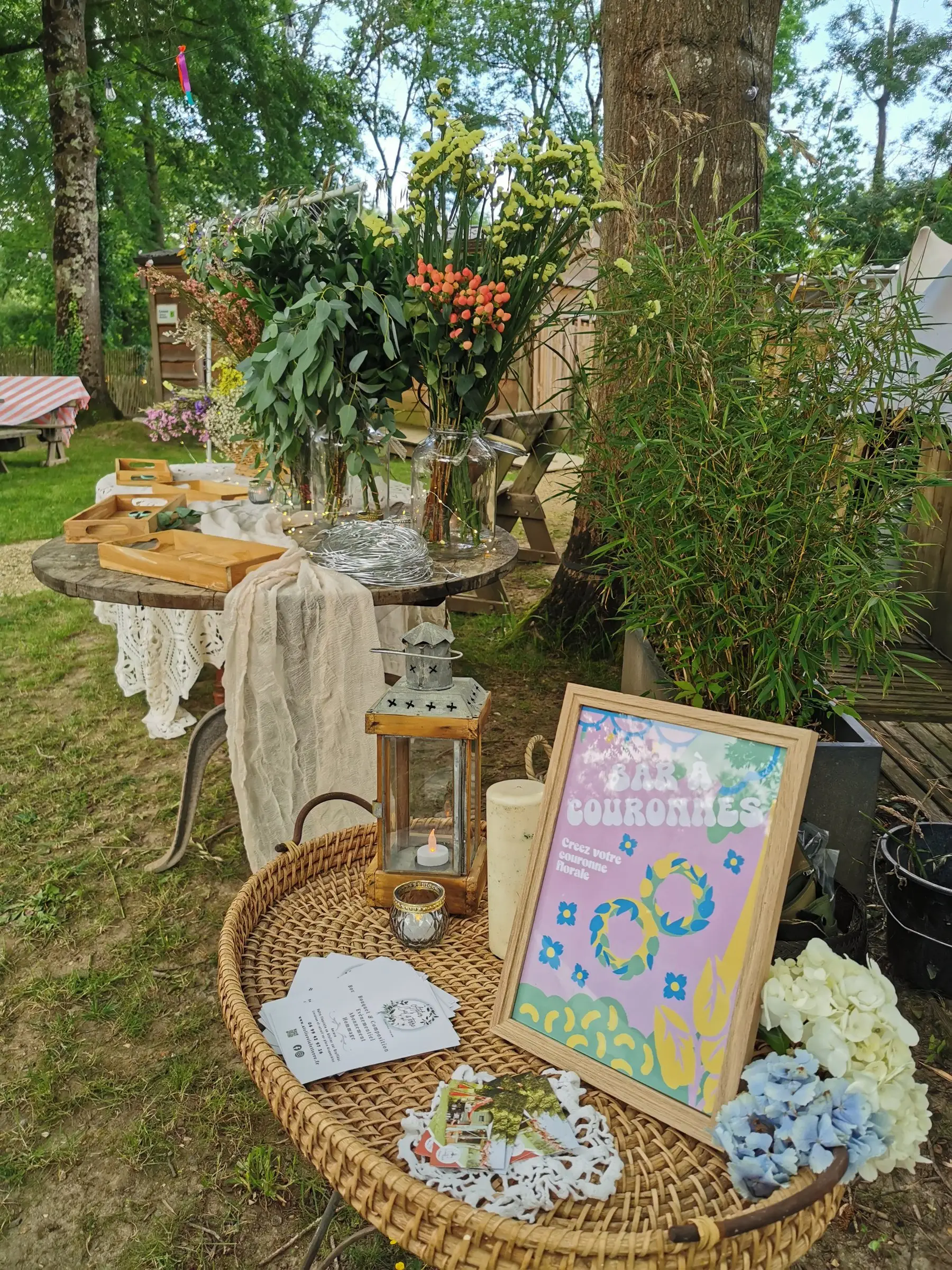 Atelier couronnes installé dans un jardin : fleurs, lanterne et panneau d’accueil.