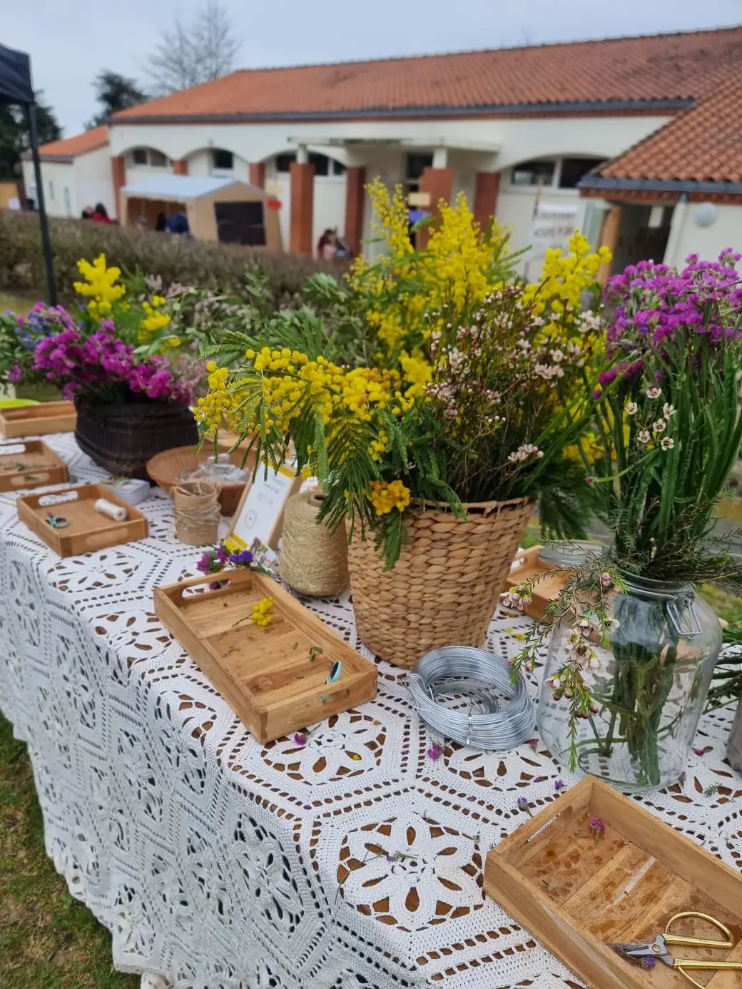 Table d’atelier dressée en plein air : nappe en dentelle, paniers de mimosa et plateaux.